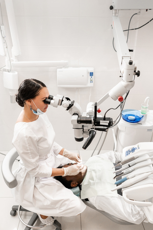 Modern equipment microscope in dental office. Young woman dentist treating root canals. Man patient lying on dentist chair with open mouth. Medicine, dentistry and health care concept.の写真素材