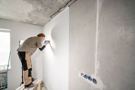 Young repairman aligns with plastic spatula. Worker glueing wallpapers on concrete wall. Repair the apartment. Home renovation concept. White Wallpaper for paintの写真素材