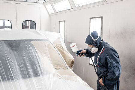 Car service station. Worker painting a white car in special garage, wearing costume and protective...