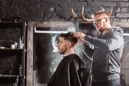 Barber sprays clean water on head in barbershop. Professional trimmer tool cuts beard and hair on young guy in barber shop salon.の写真素材