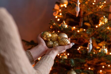 Close-up woman holds in the hands of Golden Christmas tree toys. Winter holidays in a house interior. Golden and white Christmas toys, lights garlands. Natural Danish spruceの写真素材