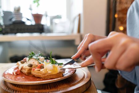 Traditional Belgian waffle with salmon, lettuce leaves and poached egg. A young woman is having Breakfast in a cafe, hands with a fork and a knife close-up.の写真素材