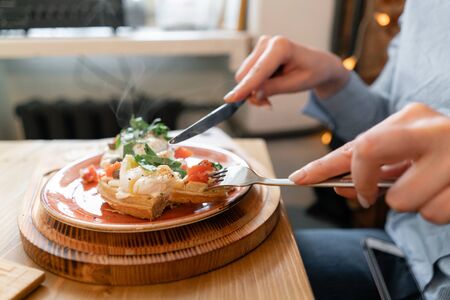 Traditional Belgian waffle with salmon, lettuce leaves and poached egg. A young woman is having Breakfast in a cafe, hands with a fork and a knife close-up.の写真素材