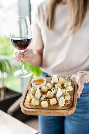 Girl holding glass red wine and wooden plate with cheese. Delicious cheese mix with walnuts, honey. Tasting dish on a wooden plate. Food for wine.の写真素材