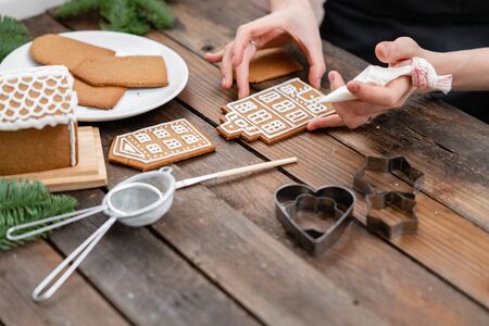 Icing of Christmas bakery. Woman decorating honey gingerbread cookies on wooden brown table....