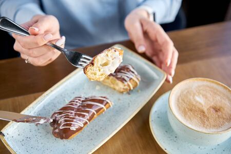 A young woman drinks coffee cappuccino in a restaurant and eats dessert. Traditional french eclairs with chocolate. Cake decorated with vanilla cream and chocolate frostingの写真素材