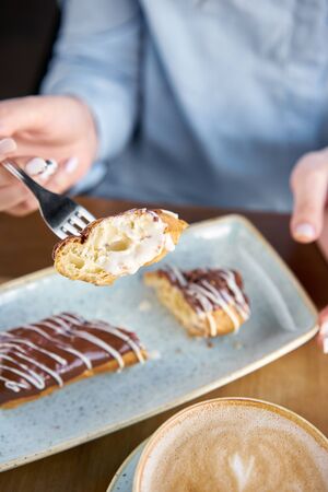 A young woman drinks coffee cappuccino in a restaurant and eats dessert. Traditional french eclairs with chocolate. Cake decorated with vanilla cream and chocolate frostingの写真素材