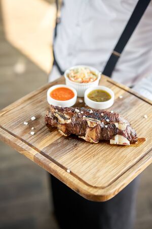 The waiter is holding a plate Beef meat cut according to the Argentine style, called asado. with Pickled cabbage and two sauces. Serving on a wooden Board.の写真素材
