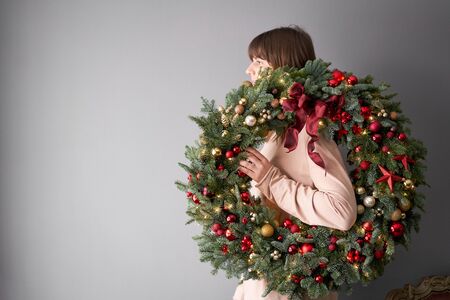 Beautiful festive wreath of fresh spruce in woman hands. Xmas circlet with red and gold ornaments and balls. Christmas mood. Gray wall on background.の写真素材