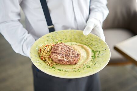 The waiter is holding a plate with Beefsteak marbled beef, served with bulgur and smoked onion...