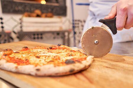 Italian kitchen and cooking concept. Chief with a pizza cutter cutting pizza to pieces at Neapolitan pizzeriaの写真素材