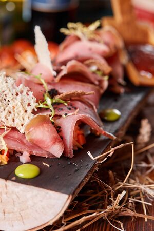 Antipasto on wooden plate close up. Cold smoked meat plate with tomato hot sauce, sliced ham, prosciutto, bacon. Appetizer on wooden tray cut tree sawed imitationの写真素材