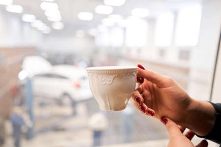 Woman drinking coffee while waiting car in the auto service station. Looking at her car sitting in waiting room of the auto service centerの写真素材