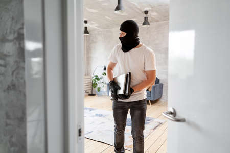 Thief with black balaclava stealing modern expensive laptop from apartment. Masked face. Man burglar stealing tv set from houseの写真素材