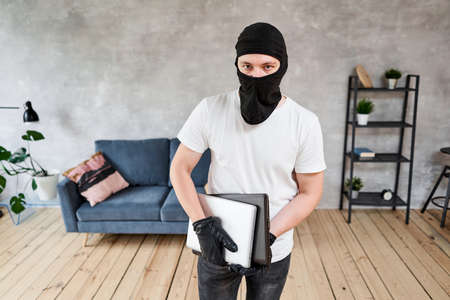 Thief with black balaclava stealing modern expensive laptop from apartment. Masked face. Man burglar stealing tv set from houseの写真素材