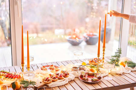 Christmas dinner feast. A small table is served with snacks, bruschettas, and canapes. A decorated dining table with champagne glasses, candles and christmas tree an garland in backgroundの写真素材
