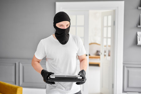 Thief with black balaclava stealing modern expensive laptop from apartment. Masked face. Man burglar stealing tv set from houseの写真素材