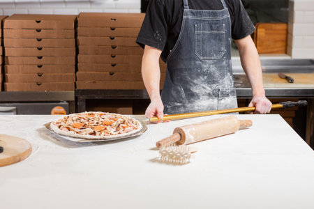 The chef prepares pizza. Raw pizza ready to bake. Cook in a blue apron in the kitchen. with a shovel...