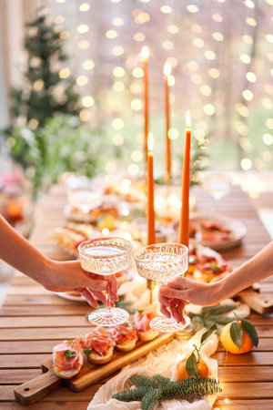 Hand with a glass of champagne in the foreground. A small table is served with snacks, bruschettas, and canapes. A decorated dining table with champagne glasses, candles and christmas tree an garlandの写真素材