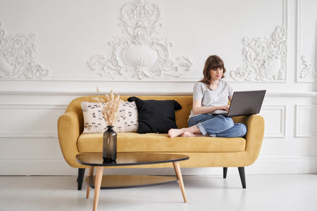 Beautiful cheerful girl using black laptop while sitting on sofa in living room at home. Young casual woman using laptop for communication and work. Stay homeの写真素材