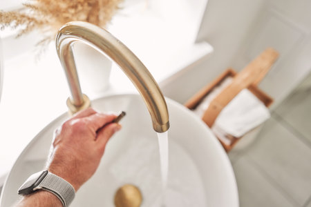 A mans hand touches the water in a beautiful sink with a golden faucet next to an oval mirror and a...