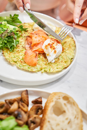 Woman cuts Zucchini pancakes, with avocado, cheese cream, salmon and egg . Healthy breakfast, protein. Restaurant dish. Breakfast in cafe on a sunny morning, on the summer verandaの写真素材