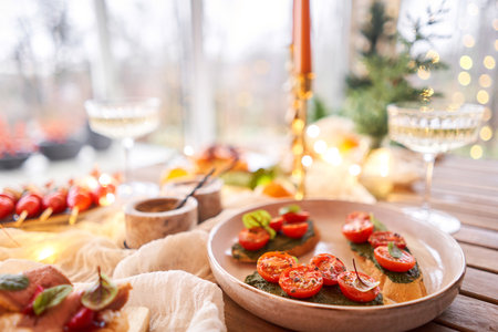 Christmas dinner feast. A small table is served with snacks, bruschettas, and canapes. A decorated dining table with champagne glasses, candles and christmas tree an garland in backgroundの写真素材
