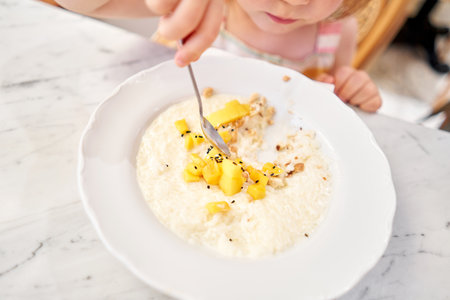 Little kid having breakfast at cafe. Adorable girl drinking still water, eating rice porridge with mango. enjoying breakfast.の写真素材