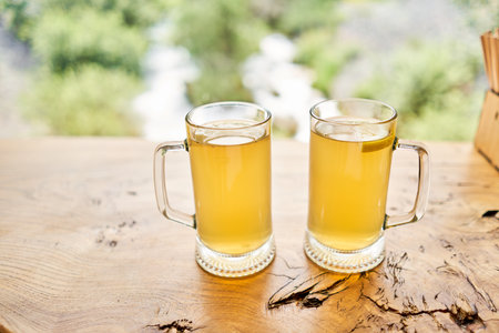 Two cups of ginger root tea with lemon, honey and mint on a wooden table and High-mountain river in the background. Walk in the mountains and relax in a cozy cafe. Hiking on a day offの写真素材