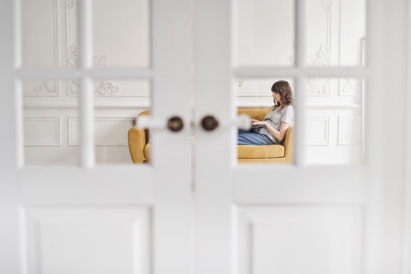 Beautiful cheerful girl using black laptop while sitting on sofa in living room at home. Young casual woman using laptop for communication and work. Stay homeの写真素材