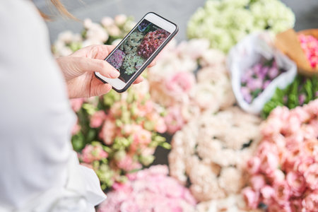 A woman takes a photo on her phone of flowers. Floral shop concept . Florist woman creates flower arrangement in a wicker basket. . Flowers delivery. A woman writes a video blog or online courseの写真素材