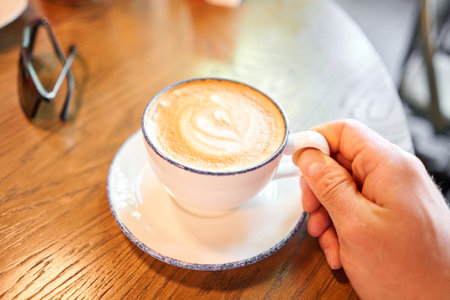 A cup of hot cappuccino on the table in the coworking space of the coffee shop. A cup of coffee on the veranda on a sunny day. Coffee break at the cafe. Close-upの写真素材