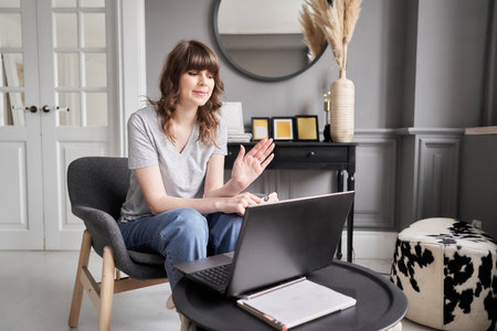 Woman waving hand has video call laptop computer online sitting in armchair at home. Hello there....