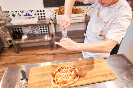 Closeup hand of chef baker in uniform blue apron cutting pizza at kitchenの写真素材