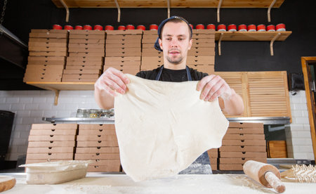 The chef prepares pizza. Raw pizza ready to bake. Cook in a blue apron in the kitchen. with a shovel in his hands. boxes for food delivery on background.の写真素材