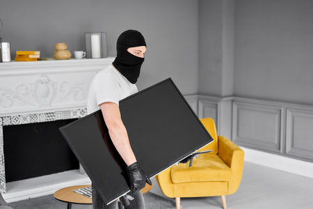 Thief with black balaclava stealing modern expensive television. Masked face. Man burglar stealing...