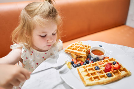 Happy 3 year old girl eat. Breakfast in cafe. Classic Viennese waffles with ice cream, berries and Maple syrup. Table in the restaurant.の写真素材