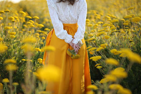 A girl in a yellow skirt and white blouse stands in a field of yellow flowersの写真素材