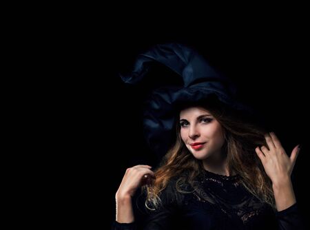 Beautiful young witch in a black hat comes out of the darkの写真素材
