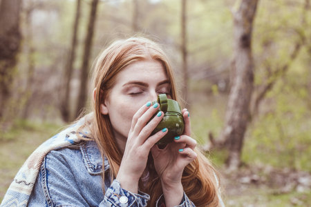 Beautiful red-haired girl wrapped in a plaid with a cup of cocoa in nature