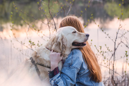 Red-haired girl hugging her dog on the river bank looking at the sunset