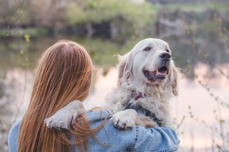Red-haired girl hugging her dog on the river bank looking at the sunset