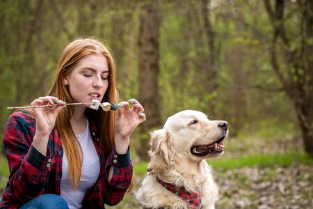 Beautiful red-haired young woman with a dog fries marshmallows on a fireの写真素材