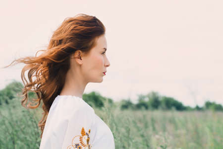 Red-haired, beautiful, young woman in a Ukrainian embroidered shirt walks in the fieldの写真素材