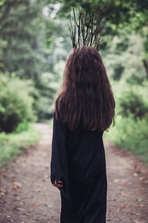 A young woman in a black dress and a crown of branches walks in the dark of the forest, back view....