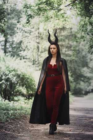 An incredibly beautiful witch in a black cloak with horns Maleficent walks through the dense forest....