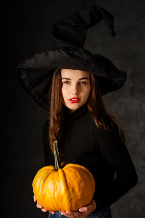 Young beautiful woman in a black witch's hat with pumpkin on a black backgroundの写真素材