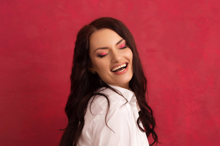Beautiful young long-haired brunette in a white shirt with a beautiful open smile on a pink backgroundの写真素材
