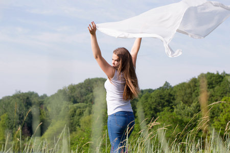 A happy young woman with a white cloth in her hands catches the wind in natureの写真素材