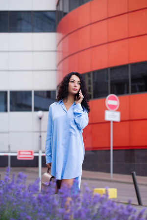 Portrait of a beautiful business woman office worker in a long blue shirt with a smartphone on the background of the cityscapeの写真素材